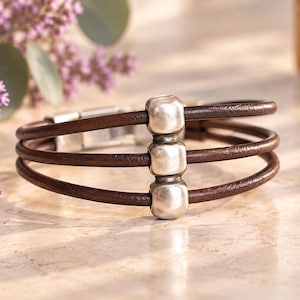 Pode incluir: Uma pulseira de várias camadas com três cordões de couro castanho escuro dispostos em paralelo. Uma fila vertical de três contas de metal prateado, lisas e arredondadas, está centrada nos cordões. A pulseira inclui um fecho metálico.