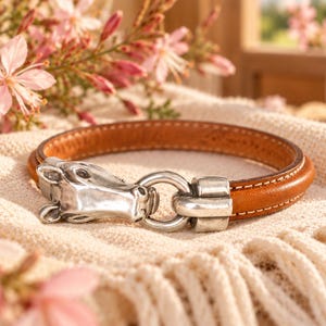 May include: A tan leather bracelet featuring a silver-toned metal horse head clasp. The bracelet has visible white stitching along the edges and a circular metal ring connector that secures the horse head to the leather band.