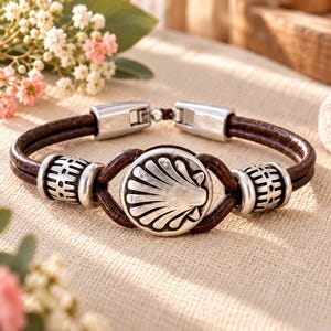 Peut inclure: Un bracelet composé de deux cordons en cuir marron foncé, d'une breloque centrale circulaire en métal argenté gravée d'un motif de coquille Saint-Jacques, et de deux perles cylindriques en métal argenté, avec un fermoir en métal argenté.