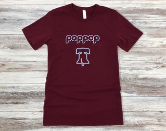May include: Burgundy t-shirt with the word "poppop" in light blue outlined text above a light blue outline of the Liberty Bell. The shirt has a crew neck and short sleeves. The shirt is displayed on a light wood grain surface.