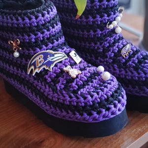 Puede incluir: Un par de zapatillas de crochet morado y negro con un logotipo de los Baltimore Ravens y otros adornos decorativos.