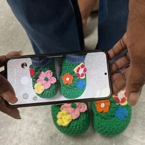 Op de afbeelding: Een paar groene gehaakte pantoffels versierd met kleurrijke, handgemaakte bloemenapplicaties. De pantoffels worden gefotografeerd, waarbij de ingewikkelde details van de bloemmotieven in roze, geel, oranje en blauw worden benadrukt.