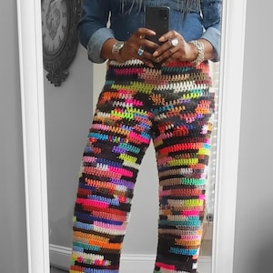 May include: A person wearing a denim shirt, a wide-brimmed hat, and colorful crocheted pants. The pants are made of different colored yarn and have a striped pattern.