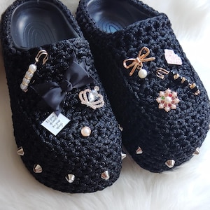 Op de afbeelding: Een paar zwarte gehaakte pantoffels met zilveren studs. De pantoffels zijn versierd met strikken, strass steentjes en andere versieringen. Een pantoffel heeft een label met de tekst "Mijn dromen zijn groter dan je vleugels".