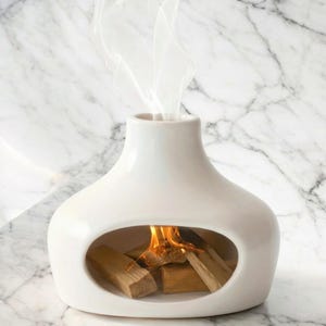 May include: A white ceramic tabletop fireplace with a rounded base and a narrow neck. The fireplace has an oval opening revealing burning wood and flames. Wisps of smoke rise from the top, creating a cozy ambiance.