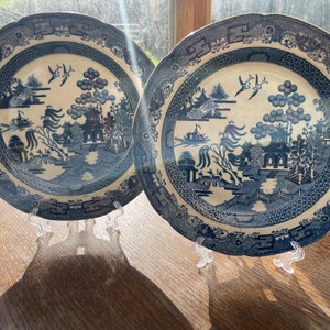 Puede incluir: Dos platos de porcelana azul y blanco con un diseño de sauce. Los platos están sobre soportes de plástico transparente.