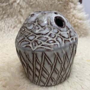 Może przedstawiać: Mały, ceramiczny wazon z teksturowanym, wzorzystym zewnętrzem. Wazon jest głównie biały z brązowymi akcentami i ciemnym otworem na górze. Projekt zawiera kombinację wzorów geometrycznych i organicznych.