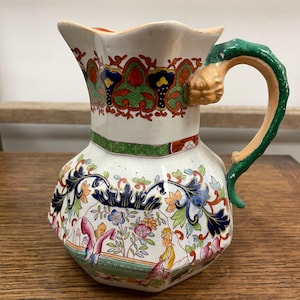 May include: A vintage white ceramic jug with a detailed floral and figure design. The jug features a green handle with a tan accent, and is decorated with red, green, blue, and orange patterns. The jug has a unique, ornate shape.