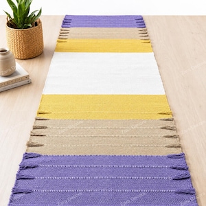 Puede incluir: Una alfombra de pasillo rectangular tejida con franjas horizontales de colores en violeta, beige, amarillo y blanco. La alfombra presenta un tejido texturizado y borlas decorativas a lo largo de los bordes largos, creando un diseño simétrico.