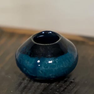 Puede incluir: Un pequeño jarrón de cerámica redondo con interior azul oscuro y exterior de dos tonos. La parte superior es negra, que pasa a un azul turquesa vibrante en la base. El jarrón está sobre una superficie de madera oscura.