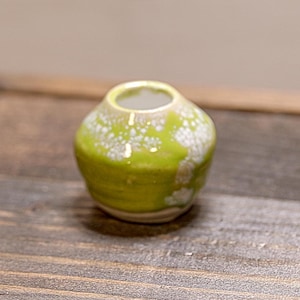 May include: A small, round ceramic vase featuring a vibrant lime green glaze with white speckled accents. The vase has a narrow, circular opening at the top and a smooth, rounded body that tapers toward the base, showcasing a glossy finish.