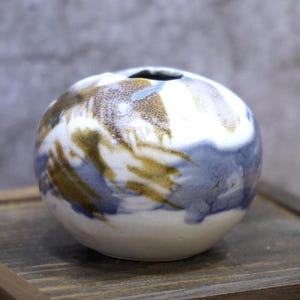 May include: A small, round ceramic vase with a narrow opening. The vase features a white base with a marbled design of blue, brown, and gold hues. The overall aesthetic is organic and artistic, suitable for displaying small flowers or as a decorative piece.