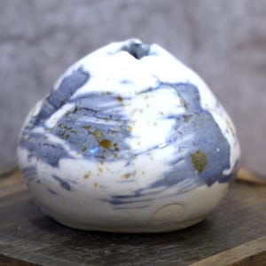 May include: A handcrafted ceramic vase with a unique, organic shape. The vase features a white base with blue and gold-toned accents, creating a marbled effect. The opening is small, and the overall design is artistic and decorative.