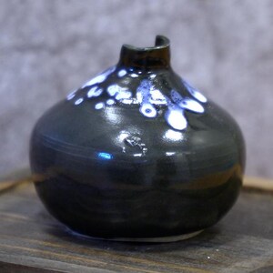 Peut inclure: Un vase en céramique sombre et brillant, avec un corps arrondi et une ouverture étroite et incurvée. Le vase est principalement noir avec des accents mouchetés blancs et bleus près du sommet. Le vase repose sur une surface en bois.