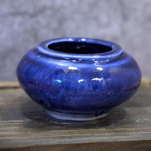 Op de afbeelding: Een kleine, ronde keramische pot met een diepblauwe glazuur. De pot heeft een brede opening en een licht uitlopende rand. De glazuur heeft kleurvariaties, waardoor een textuur ontstaat. De pot staat op een houten oppervlak.