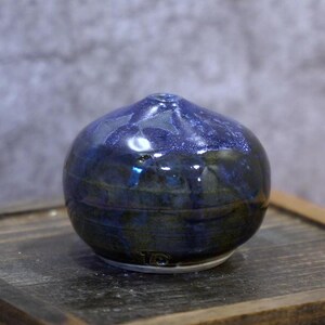 Puede incluir: Un pequeño jarrón de cerámica redondo con un esmalte azul oscuro y verde. El jarrón tiene una pequeña abertura en la parte superior y se asienta sobre una base blanca. El esmalte tiene un acabado brillante, reflejando la luz.