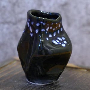Könnte beinhalten: Eine dunkelgrüne Keramikvase mit glänzendem Finish. Die Vase hat eine unregelmäßige Form mit einer leicht verdrehten Oberseite und ist mit weißen und blauen gesprenkelten Akzenten verziert. Die Vase steht auf einer Holzoberfläche.