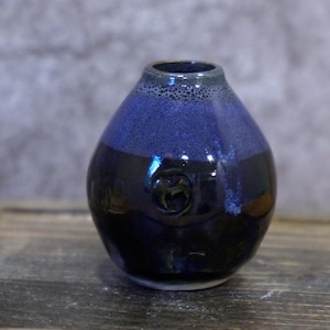 Könnte beinhalten: Eine handgefertigte Keramikvase mit dunkelblauer und schwarzer Glasur. Die Vase hat eine abgerundete Form mit einer strukturierten blauen Oberseite und einem kleinen geprägten Design auf der Vorderseite. Geeignet für die Inneneinrichtung.