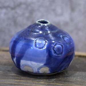 Könnte beinhalten: Eine kleine, runde, blaue Keramikvase mit einer schmalen Öffnung. Die Vase hat eine strukturierte Oberfläche mit mehreren vertieften, kreisförmigen Designs. Die Glasur hat ein glänzendes Finish mit Farbvariationen, von hell- bis dunkelblau.