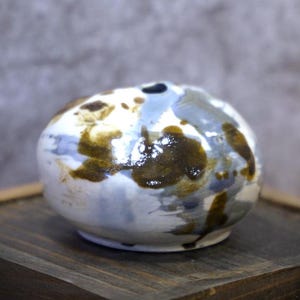 May include: A small, round ceramic vase with a unique, abstract design. The vase features a white base with brown, blue, and gray paint strokes. The vase is sitting on a wooden surface.