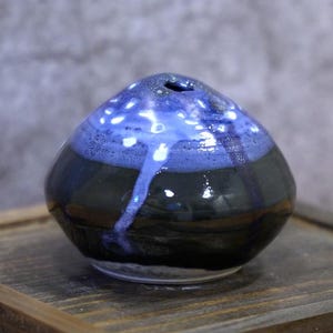 May include: A small, handcrafted ceramic vase with a rounded shape. The vase has a dark blue base, leading to a lighter blue top with white speckles. The top has a small opening. The vase sits on a wooden surface.