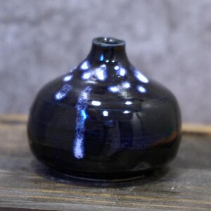 May include: A small, round, black ceramic vase with a narrow neck. The vase has a glossy finish with white and blue speckled accents. A vertical streak of white and blue glaze runs down the side.