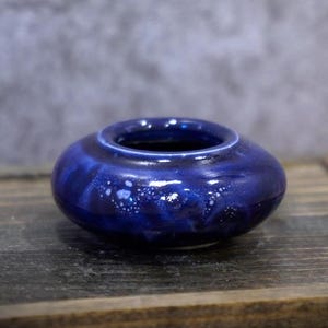 Op de afbeelding: Een kleine, ronde keramische vaas in een diepblauwe glazuur met witte spikkels. De vaas heeft een brede opening en een laag profiel, geschikt voor kleine bloemstukken of decoratieve doeleinden. De glanzende afwerking reflecteert licht.
