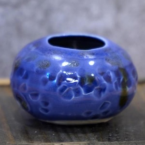 Puede incluir: Un pequeño jarrón redondo de cerámica azul con una superficie texturizada y un interior oscuro. El jarrón tiene un acabado brillante y está salpicado de detalles en azul oscuro y marrón. La abertura es circular.