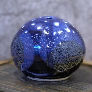May include: A spherical ceramic vase with a dark blue glaze, speckled with white and black spots. The vase has a small opening at the top and sits on a wooden surface. The glaze has a glossy finish.