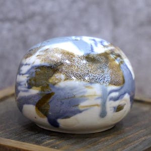 Könnte beinhalten: Eine handgefertigte Keramikschale mit runder Form. Die Schale hat eine weiße Basis mit abstrakten Mustern in Blau, Braun und Beige. Die Glasur hat ein glänzendes Finish. Die Schale steht auf einer Holzoberfläche.