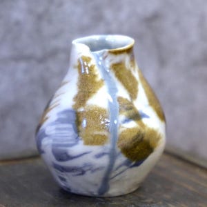 Puede incluir: Jarra de cerámica artesanal pequeña con un diseño único. La jarra presenta una base blanca con patrones abstractos marrones y azules. Una línea azul claro recorre la parte delantera. La jarra tiene un pequeño pico y una superficie ligeramente texturizada.