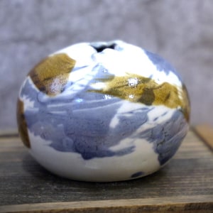 Puede incluir: Un pequeño jarrón redondo de cerámica con una abertura estrecha. El jarrón presenta una base blanca con patrones abstractos azules y marrones. El diseño recuerda a una pintura de acuarela. El jarrón está sobre una superficie de madera.
