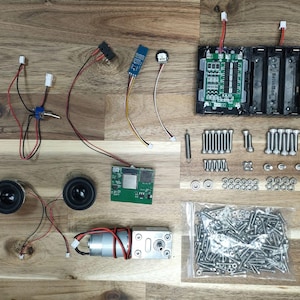 May include: An assortment of electronic components, including speakers, circuit boards, a motor, and a battery holder, are arranged on a wooden surface. Various wires, screws, and small parts are also present, suggesting a DIY electronics project or kit.