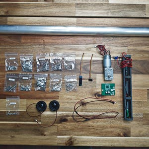 May include: An assortment of electronic components and hardware arranged on a wooden surface. Includes a long metal tube, bags of screws and bolts, a motor, circuit board, speakers, and wiring. The items are neatly organized, suggesting a DIY project or kit.
