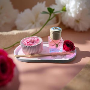 May include: A light pink oval tray holds a round, ribbed, light pink candle with a floral top, a square glass diffuser bottle with a light wood cap and a small pearl charm, and three thin wooden reeds, accented by a single dark pink flower.