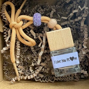 May include: A small glass bottle with a wooden top and a purple charm attached to a yellow string. The bottle has a white label with the text "I Love You Mom" and a black heart.
