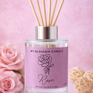 May include: A clear glass diffuser bottle with wooden reeds. The label is light purple with a rose illustration and the word "Rose". The text "My Blossom Candle" and "Natural Room Diffuser" are also visible. The background is pink.