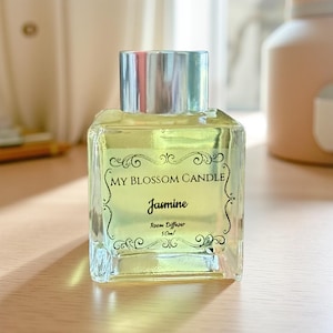 May include: A square glass room diffuser with a silver cap. The label reads "My Blossom Candle Jasmine Room Diffuser 50ml". The liquid inside is light yellow. The bottle is sitting on a light brown surface.