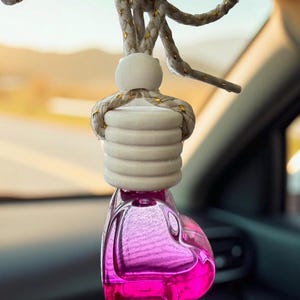 May include: A car air freshener with a heart-shaped, pink glass bottle. The bottle is topped with a white wooden piece and a braided rope with gold accents. The air freshener hangs from a black strap.