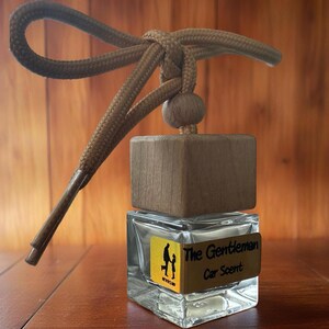 May include: A car air freshener with a clear glass bottle and a wooden top. The label reads "The Gentleman Car Scent" with a graphic of a father and child. A brown rope is attached for hanging.