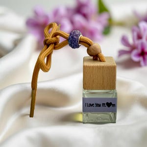 May include: A small, clear glass bottle with a wooden cap and a decorative cord. The bottle has a label that reads "I Love You Mom" with a heart symbol. The cord is mustard yellow with a blue bead and a wooden bead.
