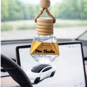 May include: Clear glass car air freshener with a wooden top and a gold label that reads "Pine Needles." The air freshener is hanging from a car's rearview mirror. The car's interior is visible in the background.
