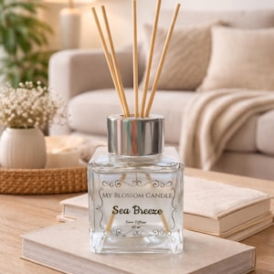 May include: A clear glass diffuser bottle with wooden reeds, labeled "My Blossom Candle Sea Breeze." The bottle has a silver cap and sits on a stack of books. The background includes a sofa, lamp, and decorative items, creating a cozy, home decor setting.