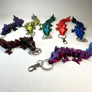 Könnte beinhalten: Eine Sammlung farbenfroher Drachen-Schlüsselanhänger in verschiedenen Farbtönen wie Lila, Rot, Grün und Blau. Jeder Drache hat ein detailliertes, stacheliges Design und ist an einem silbernen Schlüsselring oder Verschluss befestigt. Die Schlüsselanhänger sind auf einer weißen Oberfläche angeordnet.