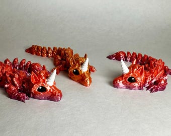 May include: Three small, 3D printed, dragon-like figurines with white horns and large eyes. The dragons are painted in shades of red and orange, with a gradient effect.