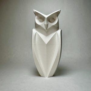 Könnte beinhalten: Eine weiße Keramik-Eulen-Skulptur mit geometrischem Design. Die Eule hat einen dreieckigen Kopf und Körper mit einem spitzen Schnabel.