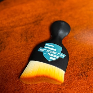 May include: A black handled brush with a logo that reads "ENGSTA RIDES" in white and blue. The brush bristles are white, transitioning to yellow and then to a reddish-orange color. The brush is set against an orange background.