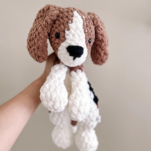 Pu&ograve; includere: Un giocattolo di peluche a uncinetto a forma di cane con pelliccia marrone, bianca e nera. Il cane &egrave; seduto con le zampe in aria.