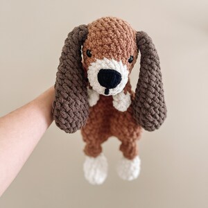 Pu&ograve; includere: Un giocattolo di peluche a forma di cane all'uncinetto con pelliccia marrone e bianca, orecchie marroni e naso nero.