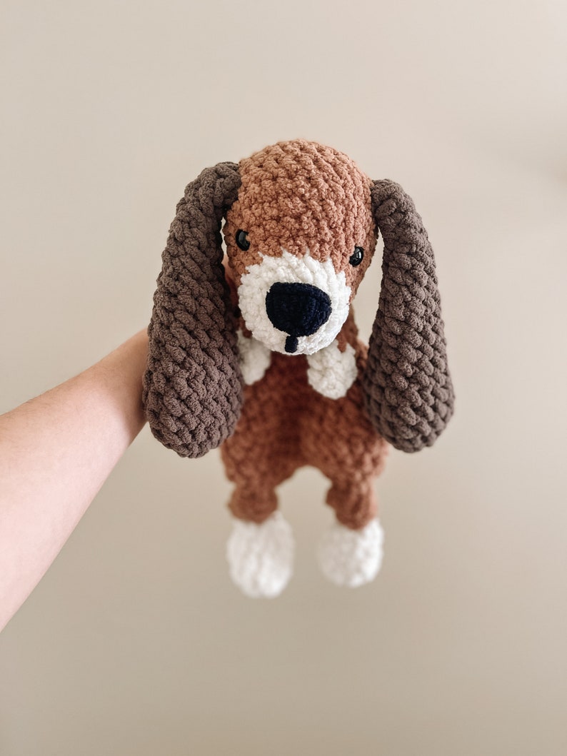 Pu&ograve; includere: Un giocattolo di peluche a forma di cane all'uncinetto con pelliccia marrone e bianca, orecchie marroni e naso nero.